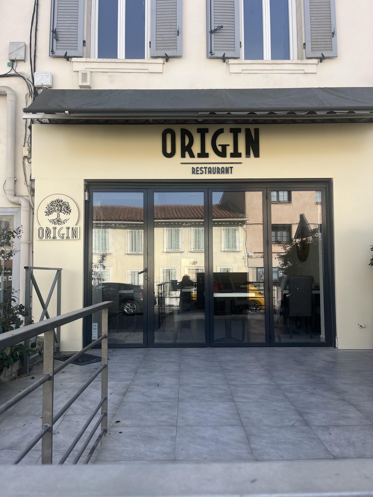 La façade du restaurant Origin : l'enseigne noire en lettres Art déco sur le mur crème, sous un store gris